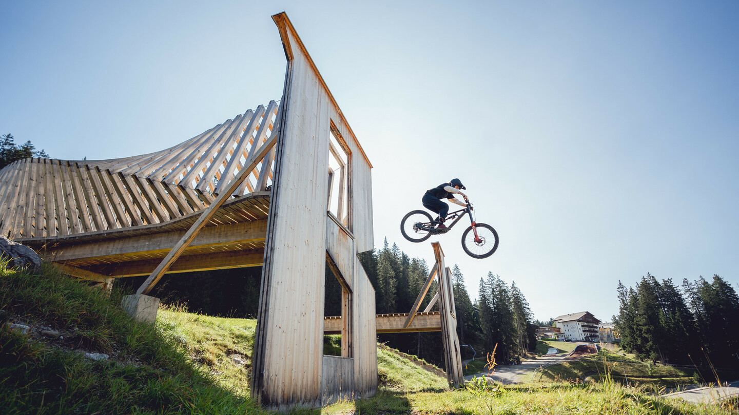 Ein Mountainbiker springt über eine Holzrampe in einem Bikepark. Die massive Holzkonstruktion hebt sich vor klarem Himmel und umliegender Natur hervor.