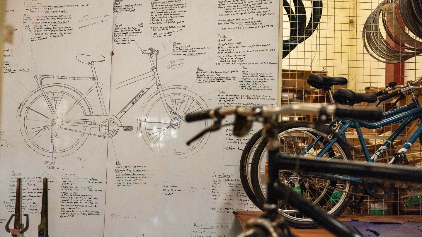 Drawing of a bicycle on a wall, surrounded by handwritten notes and technical details. Blurred bicycles and parts are visible in the foreground.