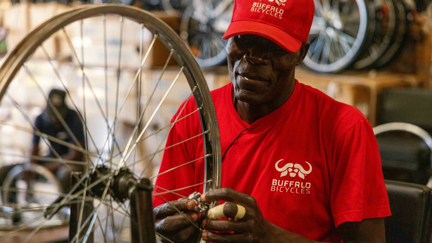 A man wearing a red shirt and cap with Buffalo Bicycles logo works on a bicycle wheel in a workshop. Stacked bicycles and boxes are blurred in the background.